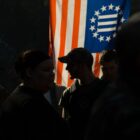 A man stands among a crowd of people in silhouette against a backlit flag of the "Three Percenter" militia.