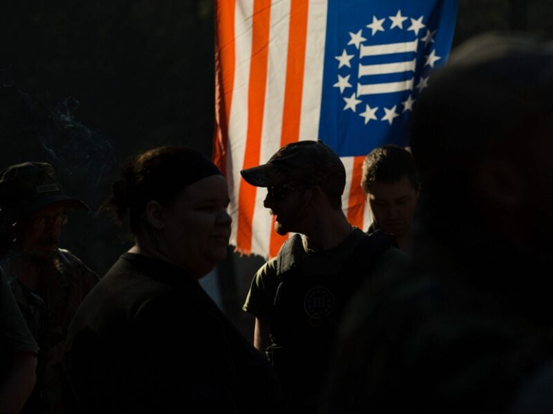 A man stands among a crowd of people in silhouette against a backlit flag of the "Three Percenter" militia.