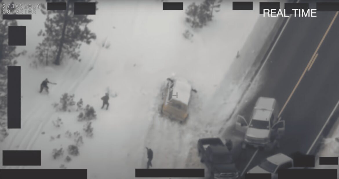 Overhead view from an FBI surveillance helicopter of several figures standing in the snow. One man is flanked by two law enforcement officers who are pointing weapons at him.
