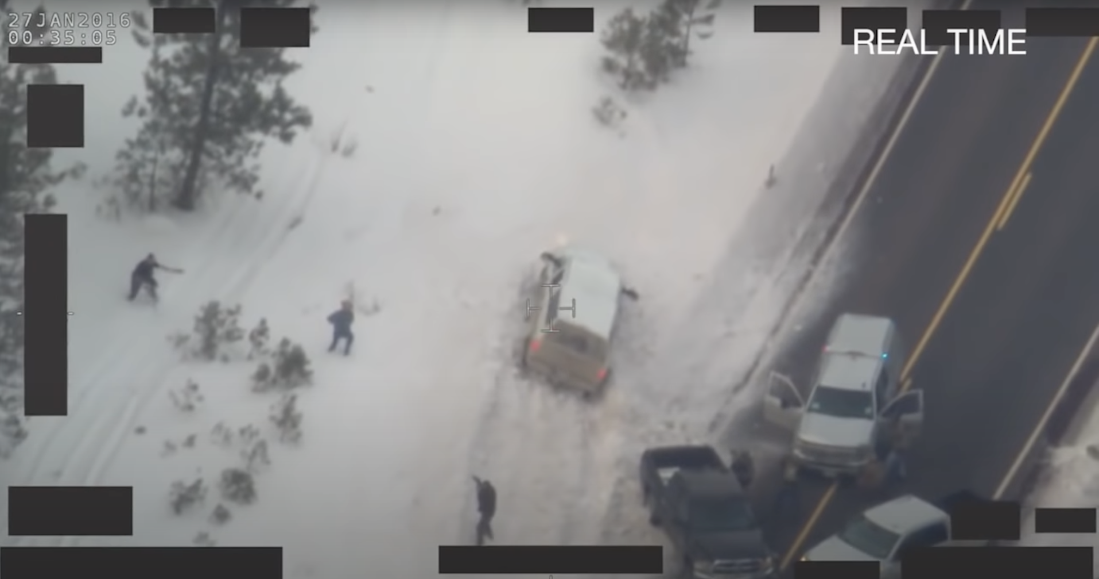 Overhead view from an FBI surveillance helicopter of several figures standing in the snow. One man is flanked by two law enforcement officers who are pointing weapons at him.