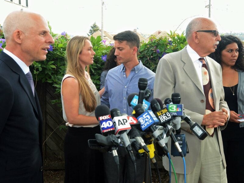 A young man and woman stand between their attorneys at an outdoor press conference.