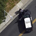 An overhead view taken from a drone shows a Black man being placed against a police SUV as he is arrested during daytime.