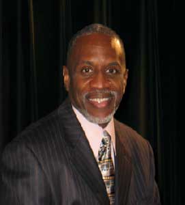 A professional portrait of Kevin H. Williams. He is a Black man in his 50s with a gray beard. He is wearing a suit.