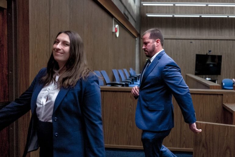 A white woman with brown, shoulder-length hair wearing a white shirt and dark blue suit smiles at the camera as she leaves a courtroom with dark wooden paneling. A ruddy 30-something male police officer with short hair, who is wearing almost the exact same color suit and a white shirt, buttons his suit jacket as he walks behind her.