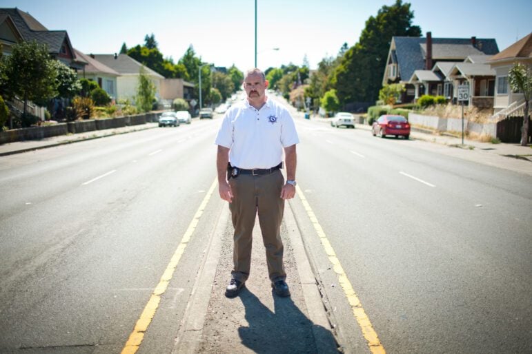 A stocky white male police officer in a white polo shirt and khakis stands facing the camera on the median of a main suburban thoroughfare during daytime. Houses from the 1800s and early 1900s frame either side of the street.