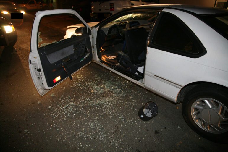 An upside-down, dark-colored baseball cap lies in a sea of auto glass next to a white sedan with its driver's side door open at night. A faint smear of blood is visible on the side of the car. The scene is illuminated by the headlights from two Vallejo police cars.
