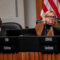 A middle-aged Black man wearing a suit sits to the right of a blonde woman who appears to be in her sixties. She is wearing glasses, a black top and a brown sweater. Both individuals are seated at a wooden city council dais. An American flag hangs behind them.