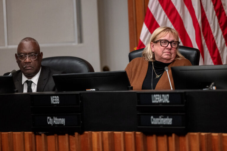 A middle-aged Black man wearing a suit sits to the right of a blonde woman who appears to be in her sixties. She is wearing glasses, a black top and a brown sweater. Both individuals are seated at a wooden city council dais. An American flag hangs behind them.