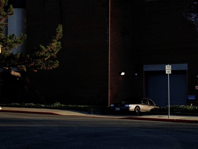 A police car sits in front of a roll-up metal door next to a "police vehicles only" sign in late afternoon light. The door is part of a large brick building. A security camera can be seen near the door.