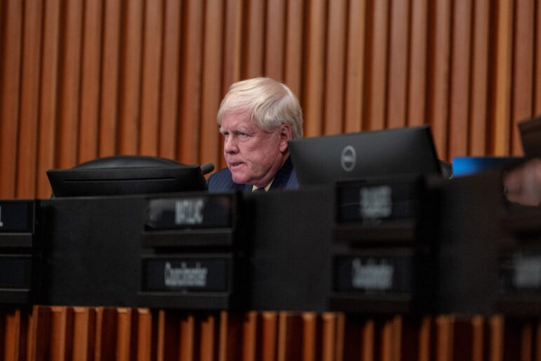 An older white man with a ruddy complexion and short white hair stares down angrily from a city council dais. A wall with warm brown wooded slats makes up the background.