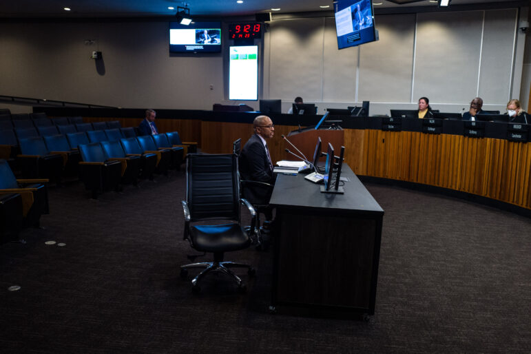 A middle-aged, professionally-dressed Black man presents to a city council from behind a black desk with computer monitors inside council chambers. The councilmembers sit on a semicircular dais. Overhead lights and computer screens hang from the walls and ceiling. Only one person is seen in the visible section of the public seating area.