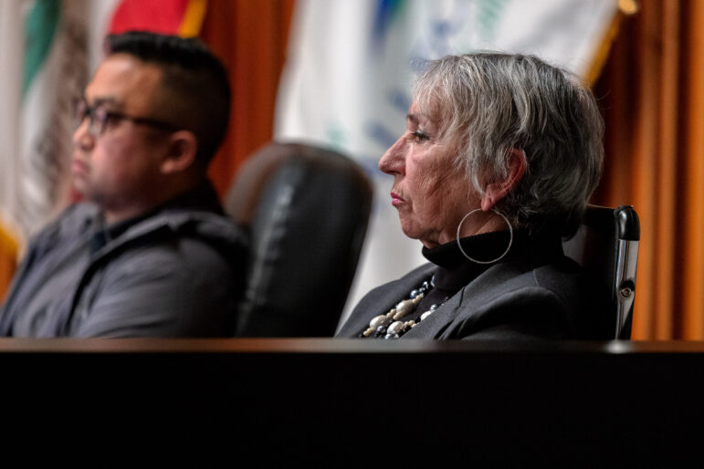 A white woman with short gray hair wearing a gray turtleneck leans back in her chair from the dais during a city council meeting.