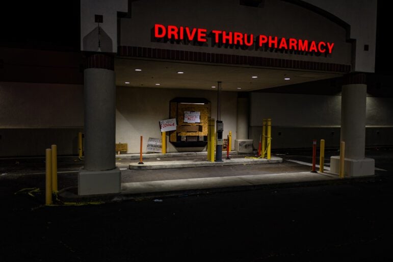 Protest signs, including signs reading "We want footage" and "Vallejo has the highest rate of people shot by police in Northern California" are lit in chiaroscuro underneath a Walgreen's "drive-thru pharmacy" sign at night. Plywood covers the pharmacy window. Crime scene tape and debris litter the ground.