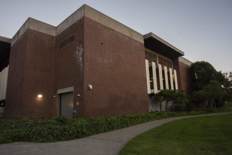A large, brick, brutalist building stands next to a park at dusk. On the left, a metal roll-up door is illuminated by a bright security light. A pan-tilt-zoom camera appears to watch the photographer.