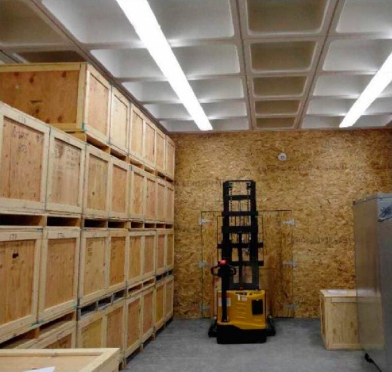 Wooden crates line the left wall of a photograph, which was made inside a storage room with a plywood back wall. Flourescent lighting illuminates the scene. A small forklift is parked near the back wall, in the center of the frame.