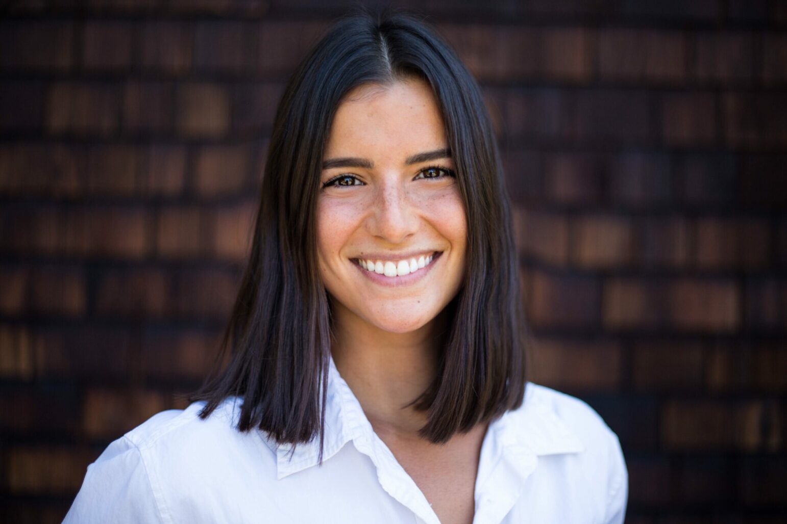 A woman in her 20s smiling broadly at the camera. She has dark shoulder-length hair and is wearing a crisp white professional shirt. She stands against a background of wood shingles that are pleasantly out of focus due to depth of field.