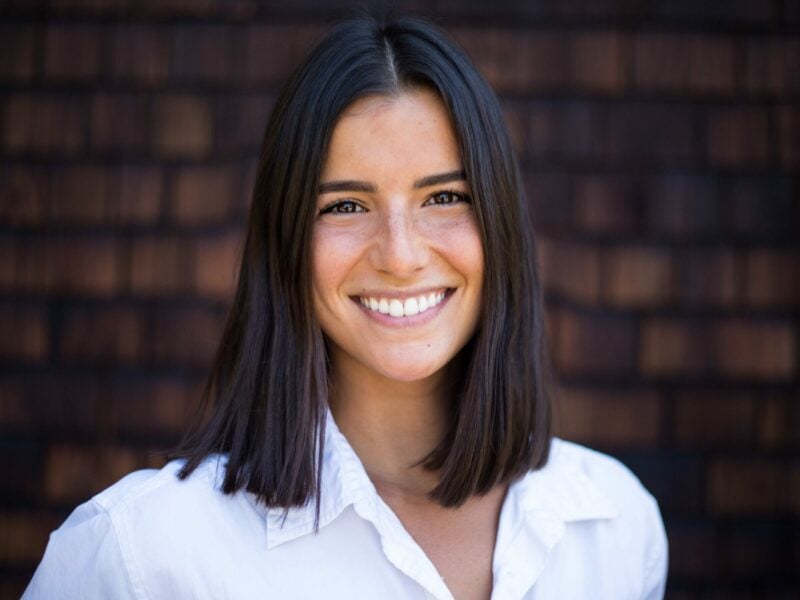 A woman in her 20s smiling broadly at the camera. She has dark shoulder-length hair and is wearing a crisp white professional shirt. She stands against a background of wood shingles that are pleasantly out of focus due to depth of field.