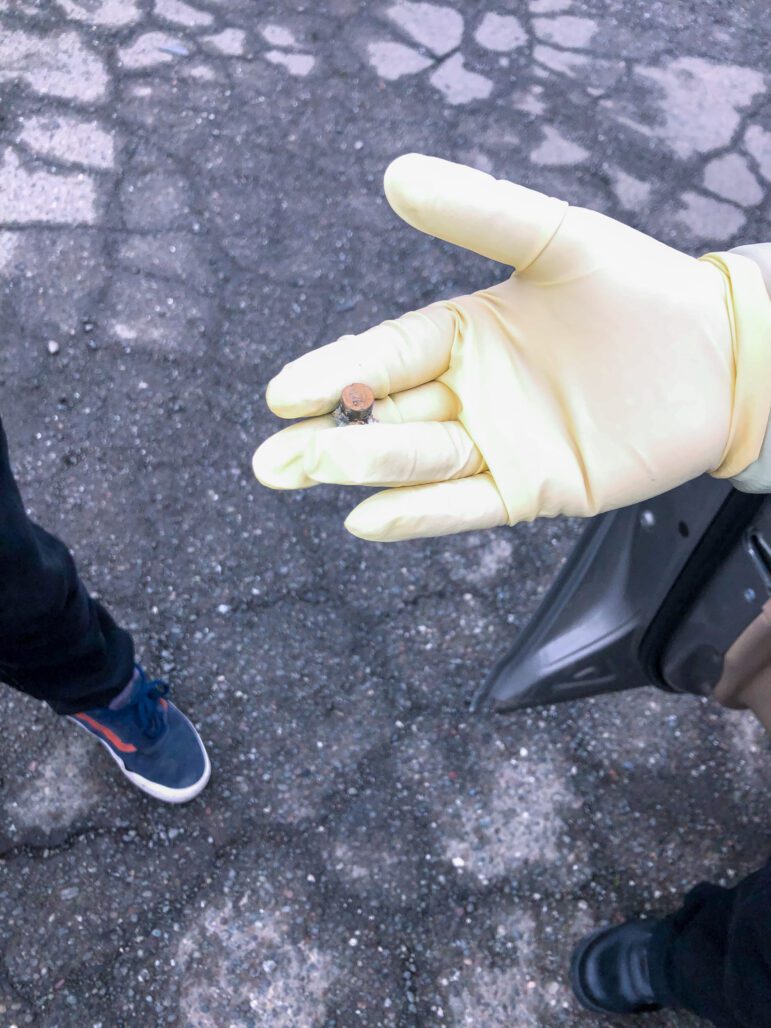 A forensic technician holds an expended bullet in their outstretched hand, which is covered by a pale surgical glove. The ground is wet, as if it has recently rained.