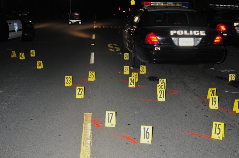 Forty-two bright yellow evidence placards mark firearm evidence scattered between two Vallejo police cars on a road at night. A third police car is visible on the right of the frame. Bright orange spray chalk or spray paint marks the ground near many of the placards.