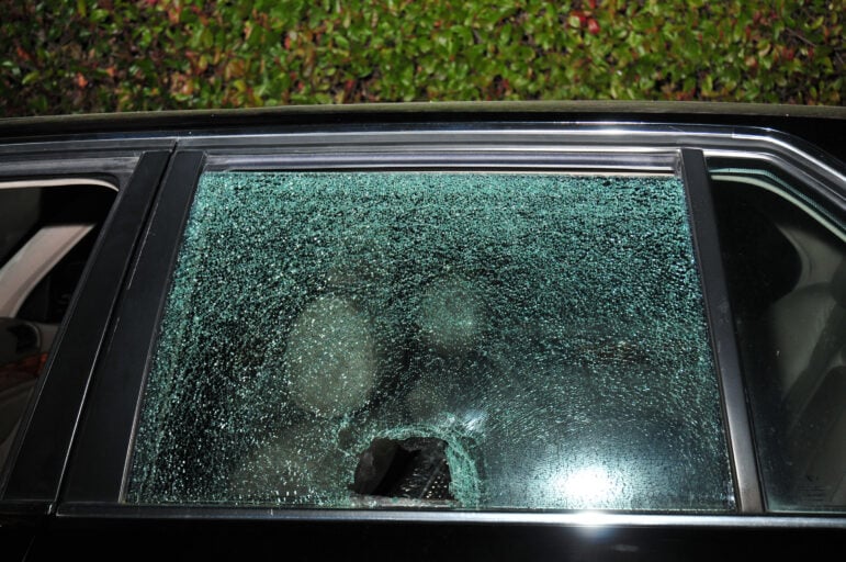An approximately five-inch diameter hole punched through the shattered rear driver's side window of a black BMW. A hint of foliage can be seen in the background. It is nighttime, and the camera flash is reflected in the window, which is fragmented but largely intact.