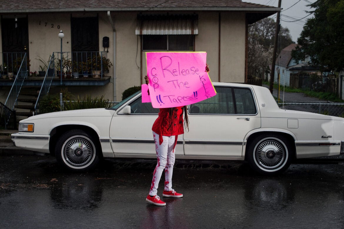 A young woman wearing a red top, red shoes and white and red pants holds a bright pink handwritten sign that reads, "Release the tapes!! Killer Cop" She stands in front of a large, off-white sedan from the 1980s or early 1990s. The background is dark and it appears to have been raining.