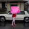 A young woman wearing a red top, red shoes and white and red pants holds a bright pink handwritten sign that reads, "Release the tapes!! Killer Cop" She stands in front of a large, off-white sedan from the 1980s or early 1990s. The background is dark and it appears to have been raining.