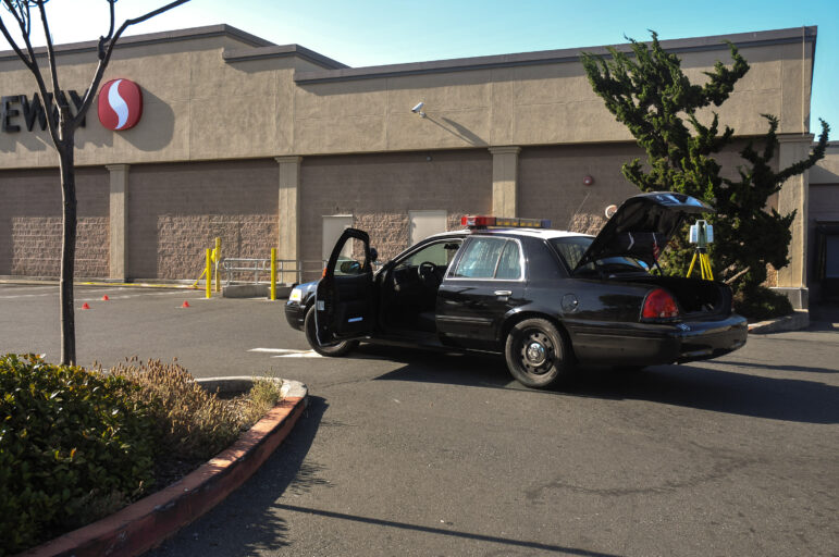 A white CCTV surveillance camera looks down on bright orange crime scene placards in the parking lot of a Safeway grocery store in daytime. A Vallejo police car is in the foreground, its driver's side door and trunk open. A crime scene measurement tool stands on a yellow tripod behind the police car.