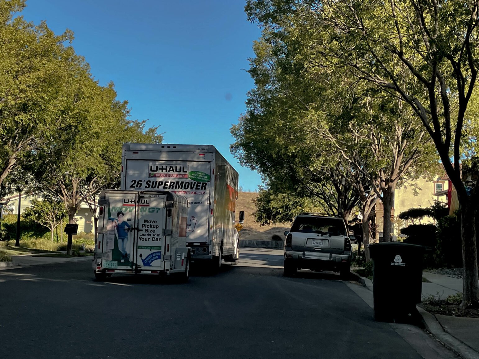 A large U-Haul moving truck labeled '26' Super Mover' is parked on a suburban street during daylight. Behind it, a smaller U-Haul trailer is attached. Trees line the street, and a few houses are visible in the background.