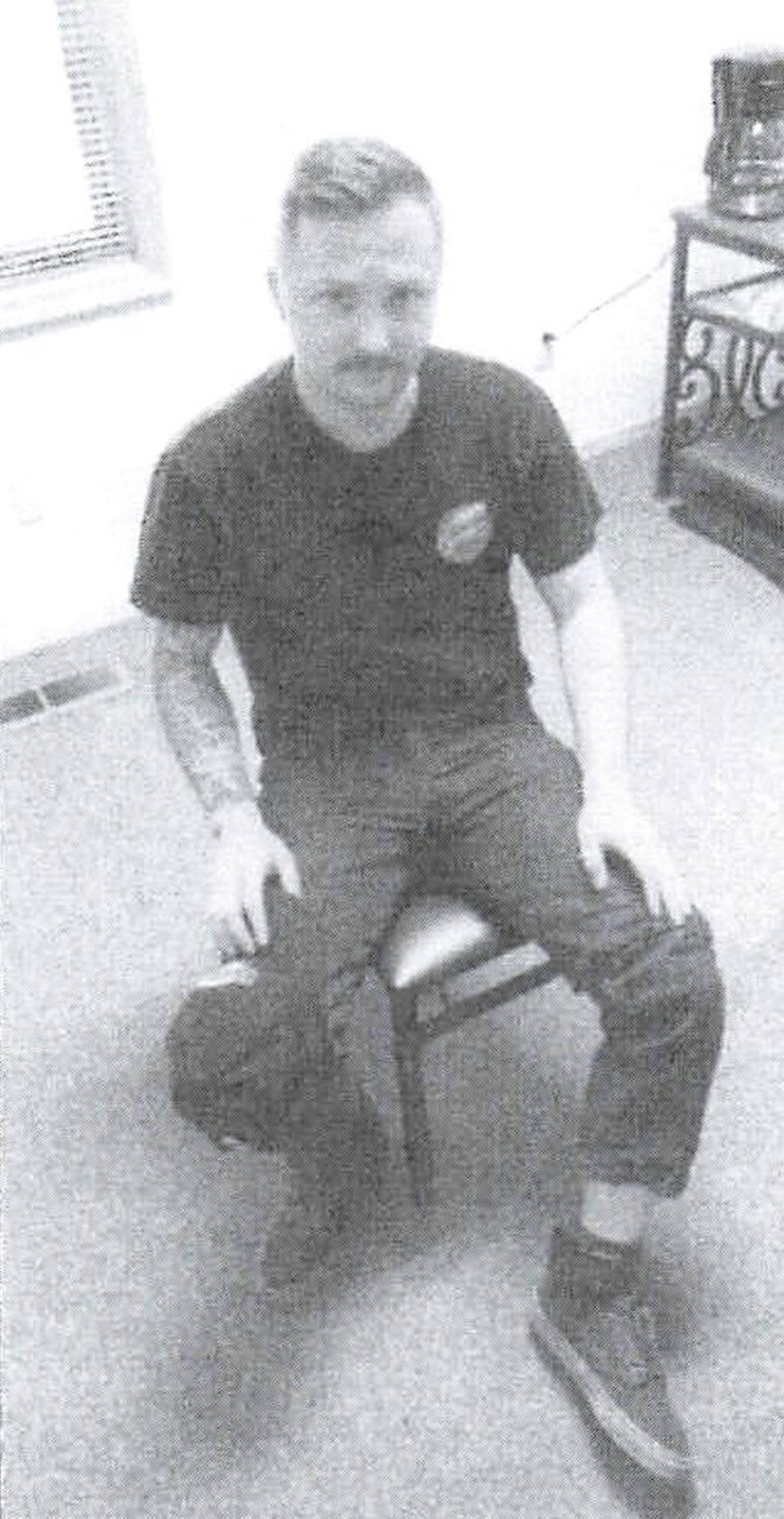 A grayscale photocopied photograph of a young man with short hair with a neutral expression as he sits on a chair in a sparsely furnished room. He is wearing a black t-shirt with a logo, and his jeans are rolled up to show an ankle monitor on his right leg.