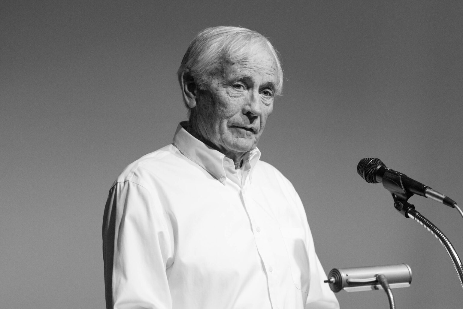 Black and white photo of a man speaking into a microphone. He is dressed in a white shirt and appears to be engaged in a serious discussion, with a thoughtful expression. The background is a soft gray, highlighting his face and the microphone in front of him.