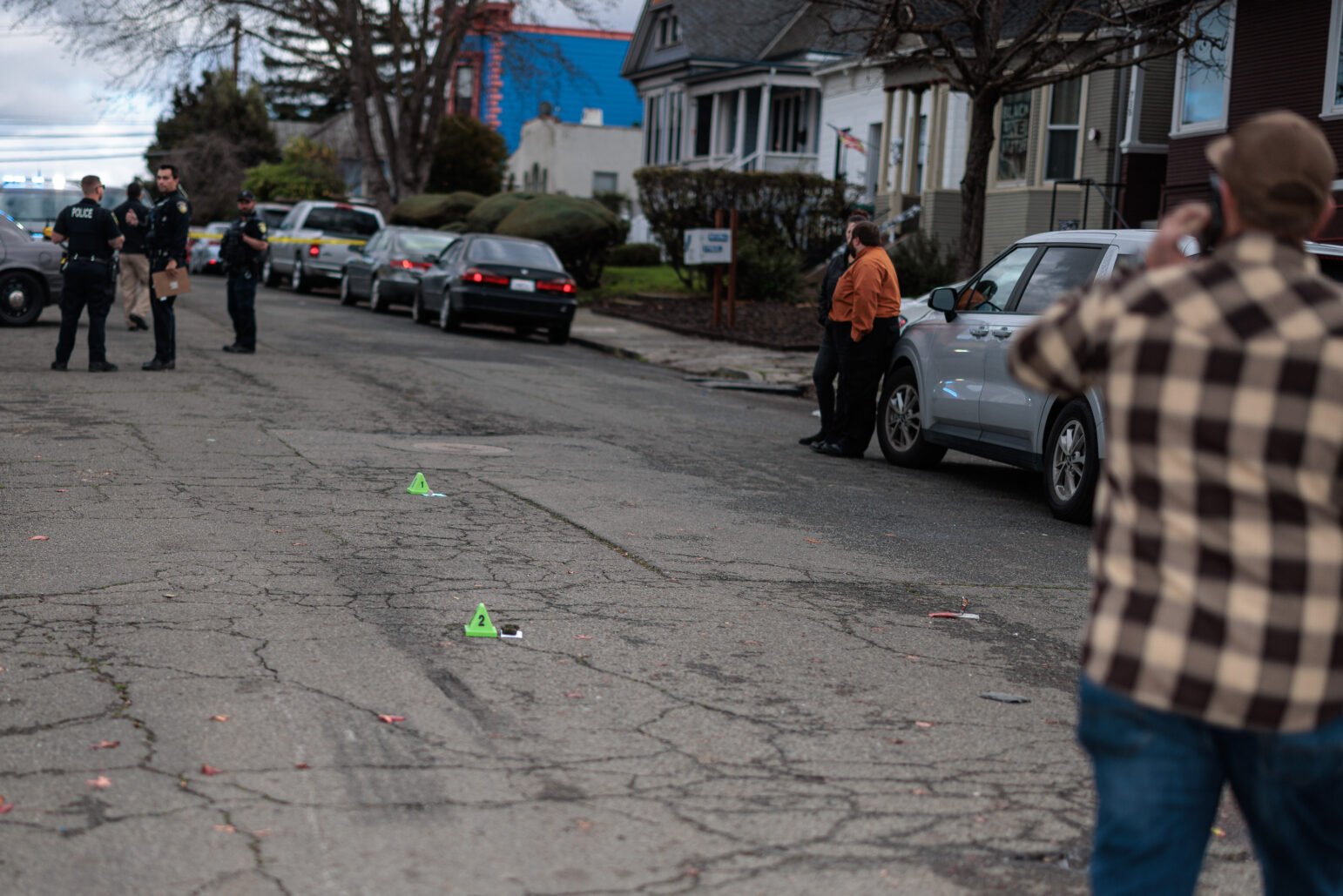 A Vallejo police detective talks on a cell phone near a group of other officers while standing in the middle of a residential street during the daytime. Two bright green evidence placards can be seen center frame.