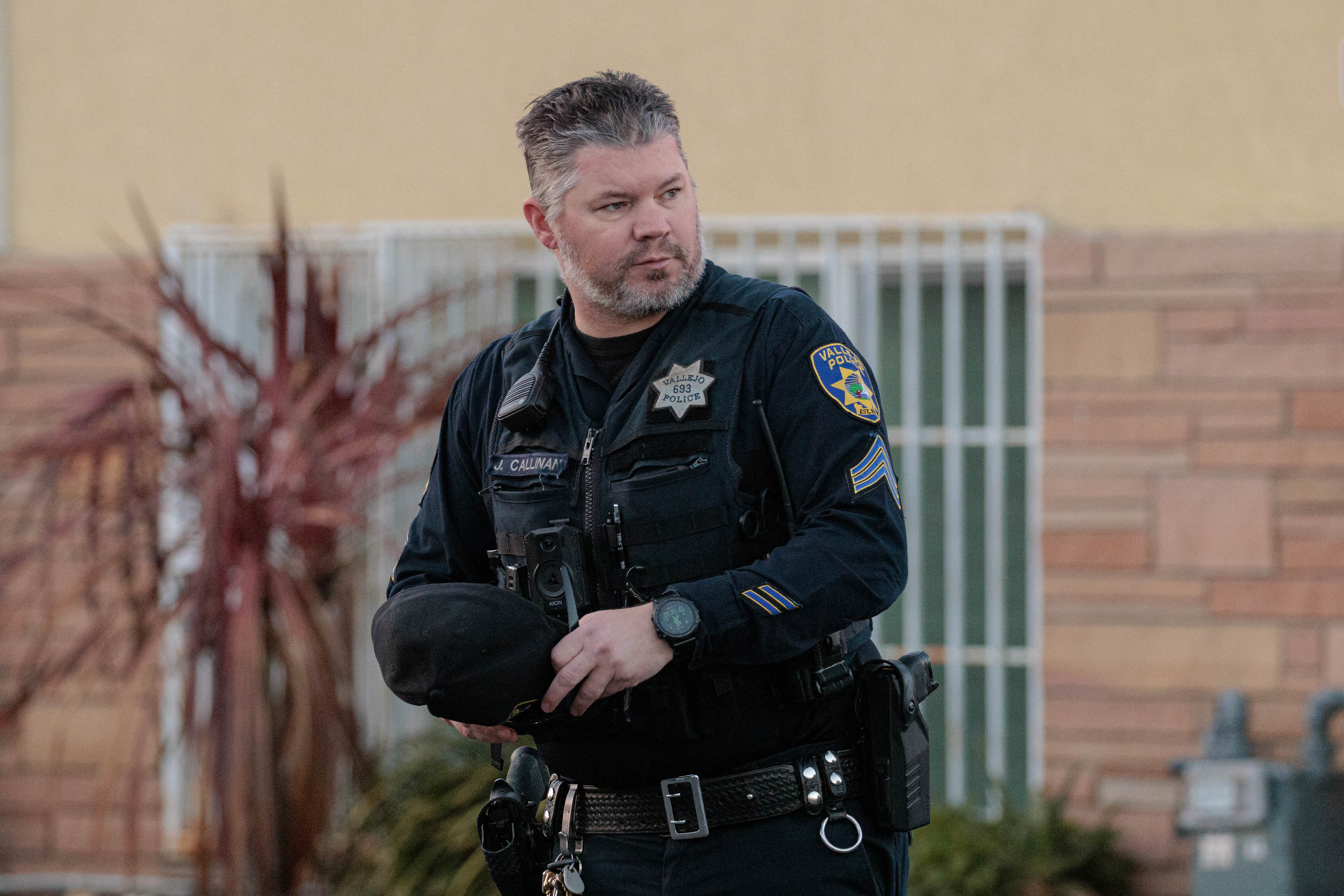 A police officer in a tactical uniform, equipped with a badge, body camera, and various gear, stands alert in an urban environment. His expression is serious and focused. The backdrop features a light-colored building and a decorative plant, emphasizing the contrast between the officer's dark uniform and the surroundings.