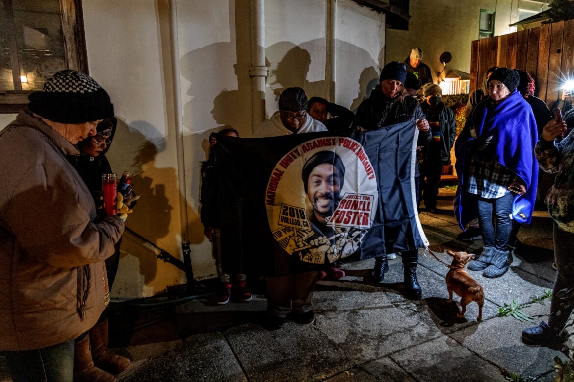 A crowd of people gathers for a vigil behind a building at nighttime. A Black woman holds a flag with the visage of Ronell Foster, a 33-year-old father of two who was killed by Vallejo police in 2018. The flag reads "National Unity Against Police Brutality," "2018 Vallejo CA," and "Justice for Ronell Foster."