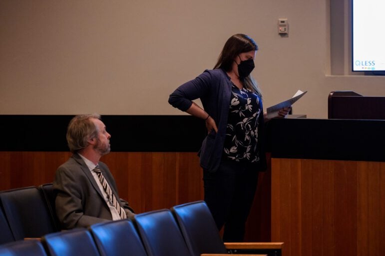 A woman in her 30s wearing a medical mask is observed reading her speaker's notes by a man in his 60s who is not wearing a mask in a city council chambers.