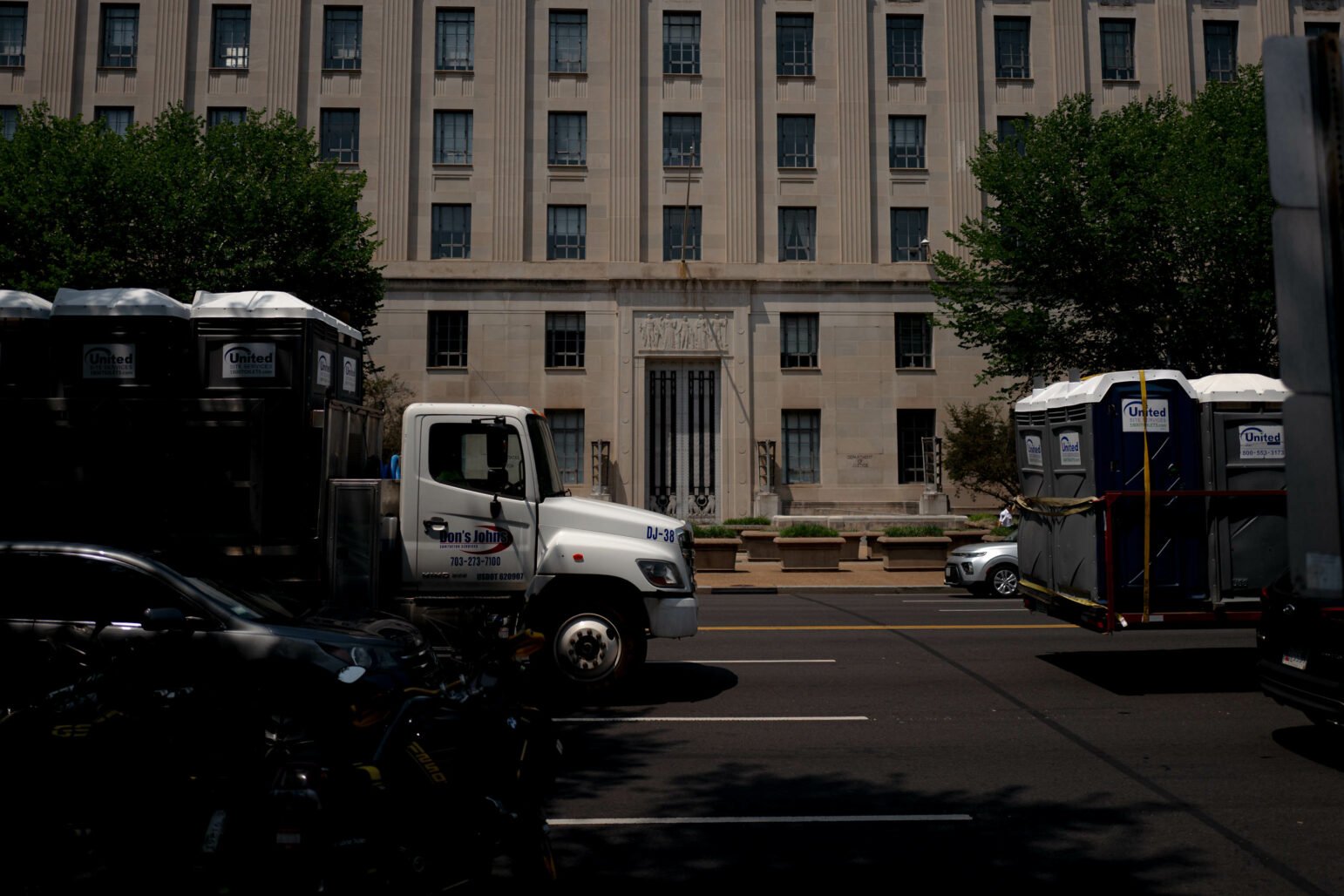 A truck labeled 'Don's Johns' parked on the street in front of the Robert F. Kennedy Department of Justice Building. The truck is carrying portable toilets. The building's facade is visible in the background with trees on either side of the street.