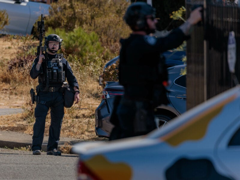 Two SWAT officers in tactical gear during a daylight operation in a suburban setting. One officer is standing while carrying a rifle, wearing a helmet and protective glasses. The other officer is standing beside a fence, pointing a handgun up toward a second-story window. The background shows dry vegetation.