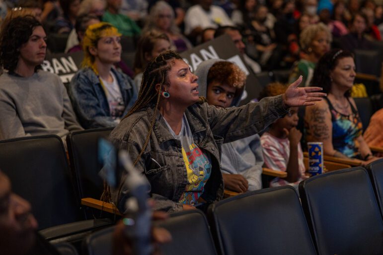 A woman in her 30s gestures with an open palm and leans forward, tears in her eyes, as she speaks to a city council. She is surrounded by community members, some of whom hold black and white signs that read, "Ban Killer Kops."