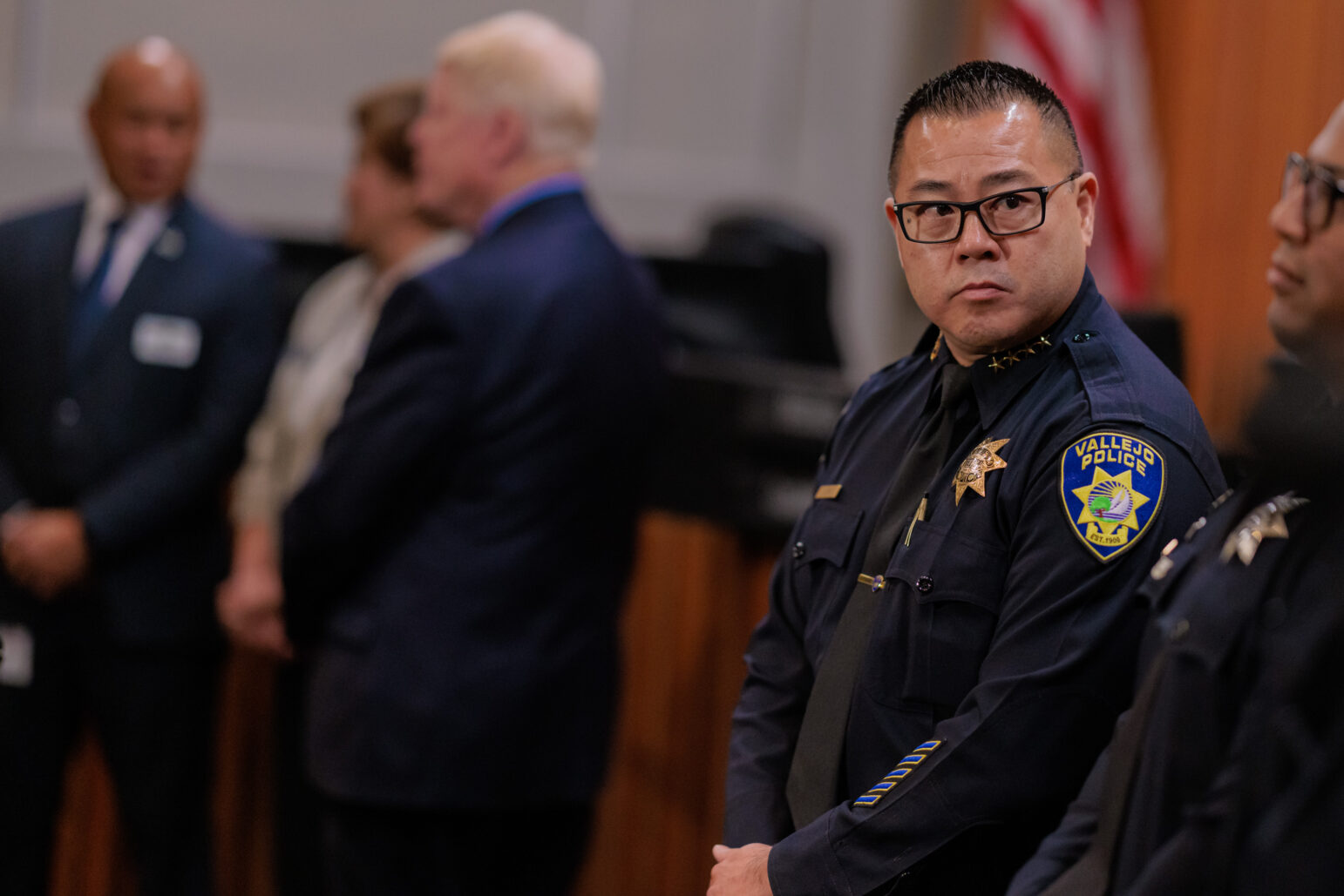 A focused police chief in uniform with the Vallejo Police badge visible, standing with his hands clasped in front of him. In the softly blurred background, several individuals are conversing, wearing professional attire. The setting is inside a city council chambers during a press conference at which California Attorney General Rob Bonta announced a lawsuit and consent decree against he Vallejo Police Department.