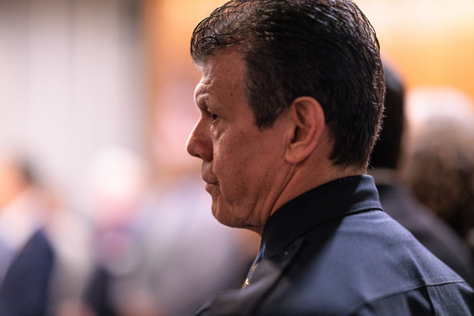 A close-up side profile of a man attending an indoor event. The focus is on his contemplative expression. He has dark, slicked-back hair and wears a dark shirt with a collar. The background is softly blurred, with indistinct figures suggesting a crowd in a large room.