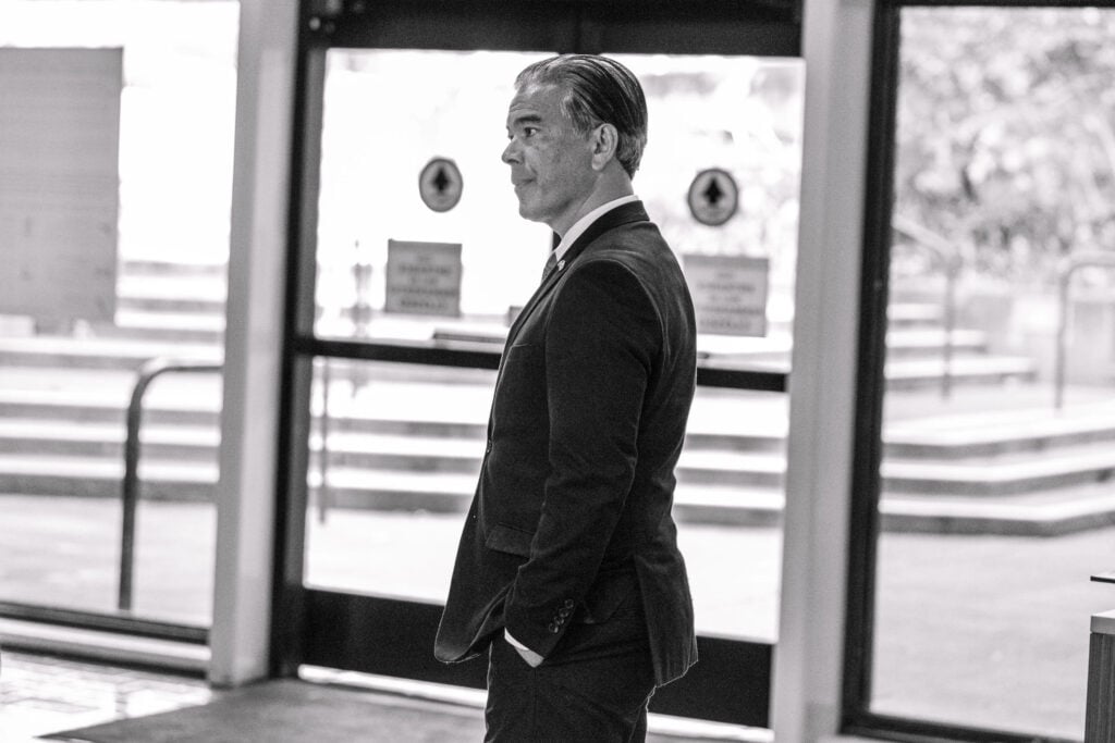 A black and white photo of California Attorney General Rob Bonta standing in profile in an indoor setting. He's wearing a dark suit and a light blue tie, with a lapel pin on his jacket. He appears focused, looking off into the distance with a pensive expression. The background is an out-of-focus view of a public building entrance, with doors and directional signs slightly visible, casting soft natural light on the subject.
