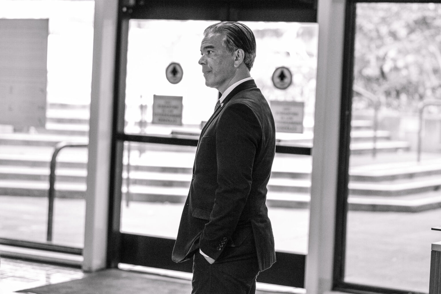 Black and white photo of California Attorney General Rob Bonta standing in profile in an indoor setting. He's wearing a dark suit and a light blue tie, with a lapel pin on his jacket. He appears focused, looking off into the distance with a pensive expression. The background is an out-of-focus view of a public building entrance, with doors and directional signs slightly visible, casting soft natural light on the subject.