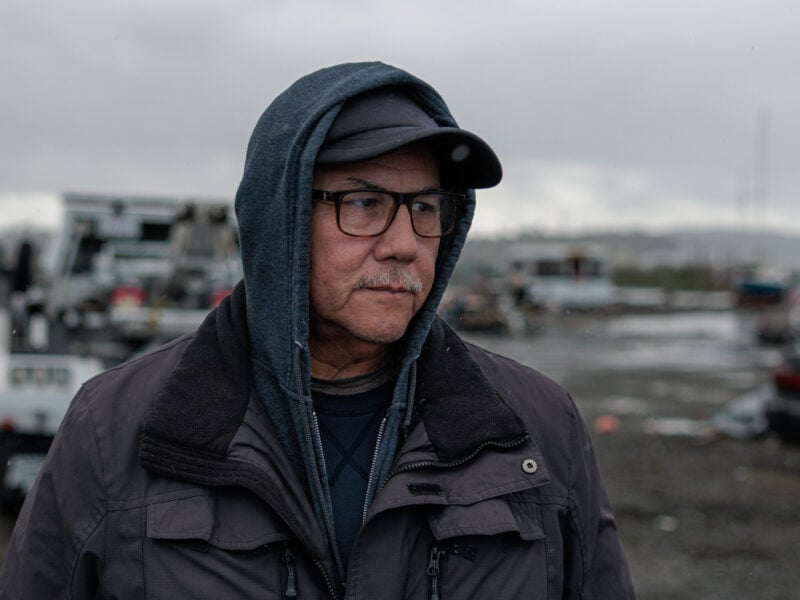 A person in a hooded jacket and glasses stands in a homeless encampment, with debris and vehicles in the background. The sky is overcast, and the ground is muddy.