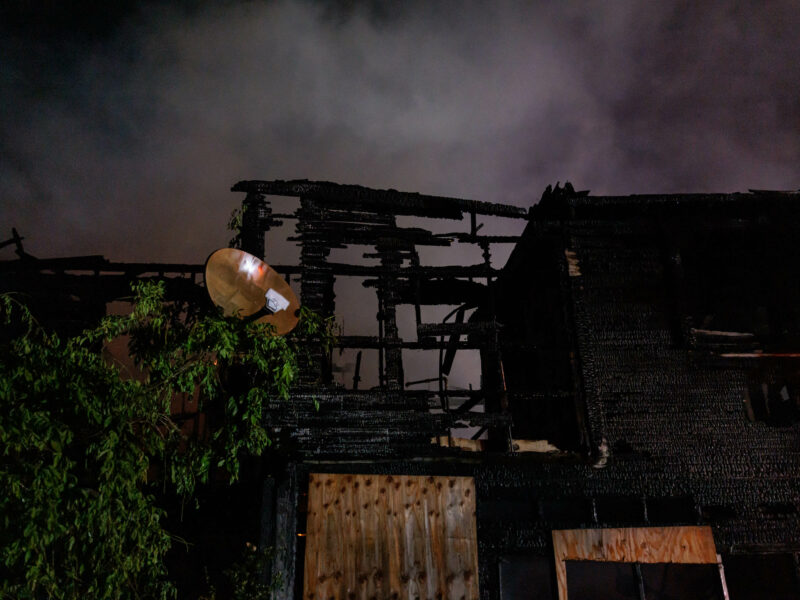An aftermath of a house fire at night, showing the charred remains of a two-story home. A satellite dish is partially visible amid the ruins, and smoke rises against a dark sky, illustrating the devastating effect of the fire.