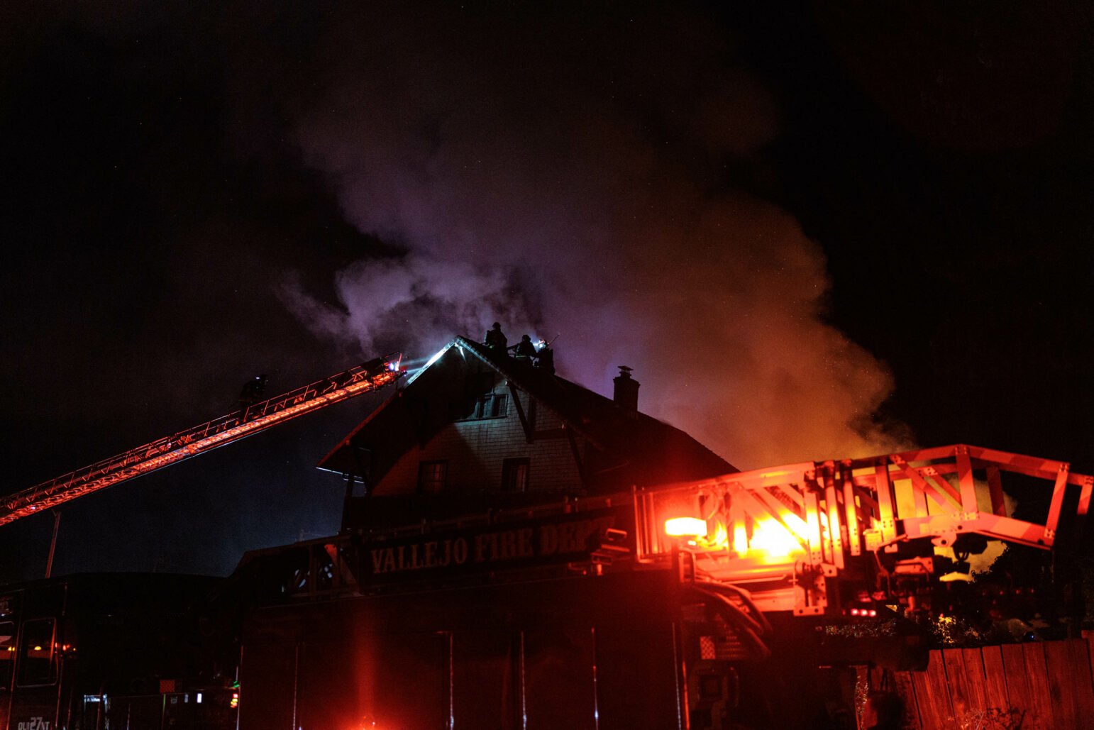 A dramatic nighttime scene showing firefighters battling a blaze at a large multi-story house. The roof of the house smokes under a dark sky, with firefighters visible on the roof via a ladder truck, working to control the fire, highlighting the intensity and danger of their work.