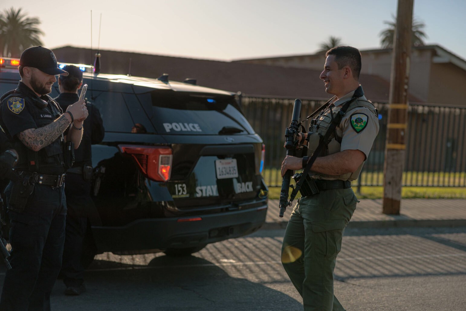 A police officer takes a photo with a phone of a sheriff's deputy holding a rifle. Both officers are in uniform, and a police SUV with flashing lights is parked behind them.