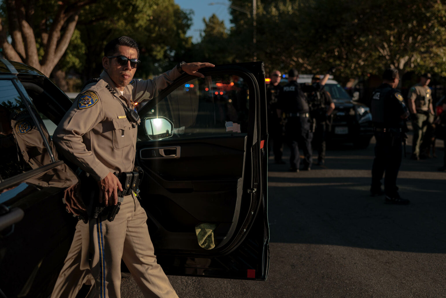 A California Highway Patrol officer in uniform leans against the open door of a patrol car. He is wearing sunglasses and looking toward the camera with a serious expression. Other officers and patrol cars with flashing lights are visible in the background, along with trees.