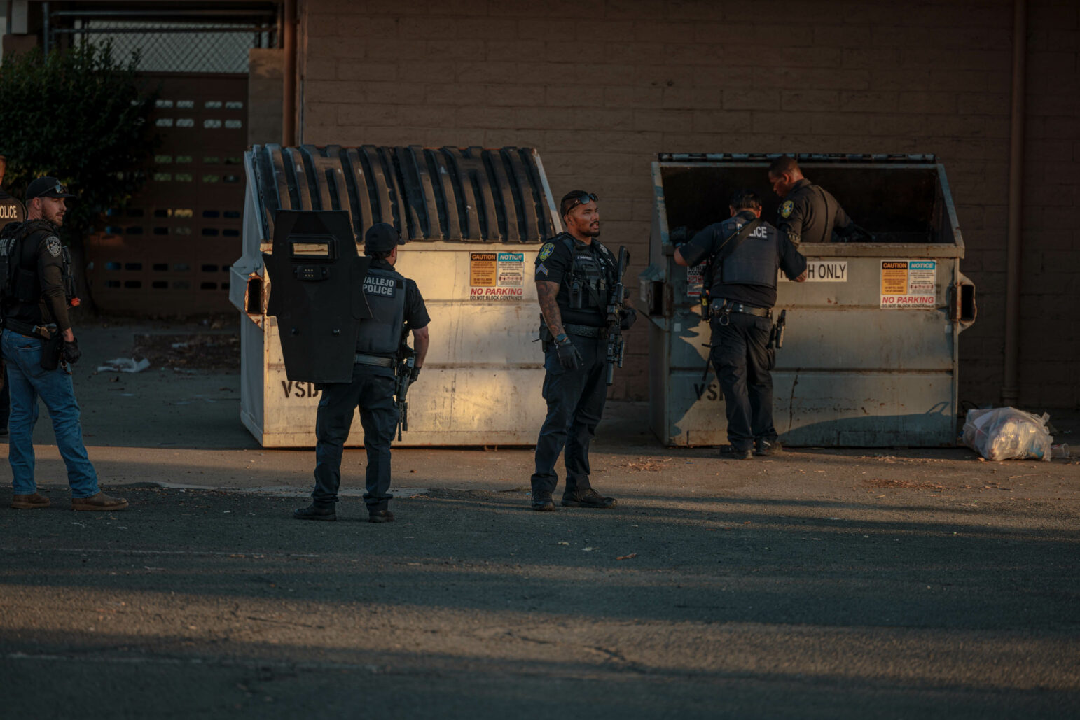 Four police officers in uniform are inspecting and searching dumpsters behind a school. One officer is holding a shield, and others are looking inside the dumpsters.