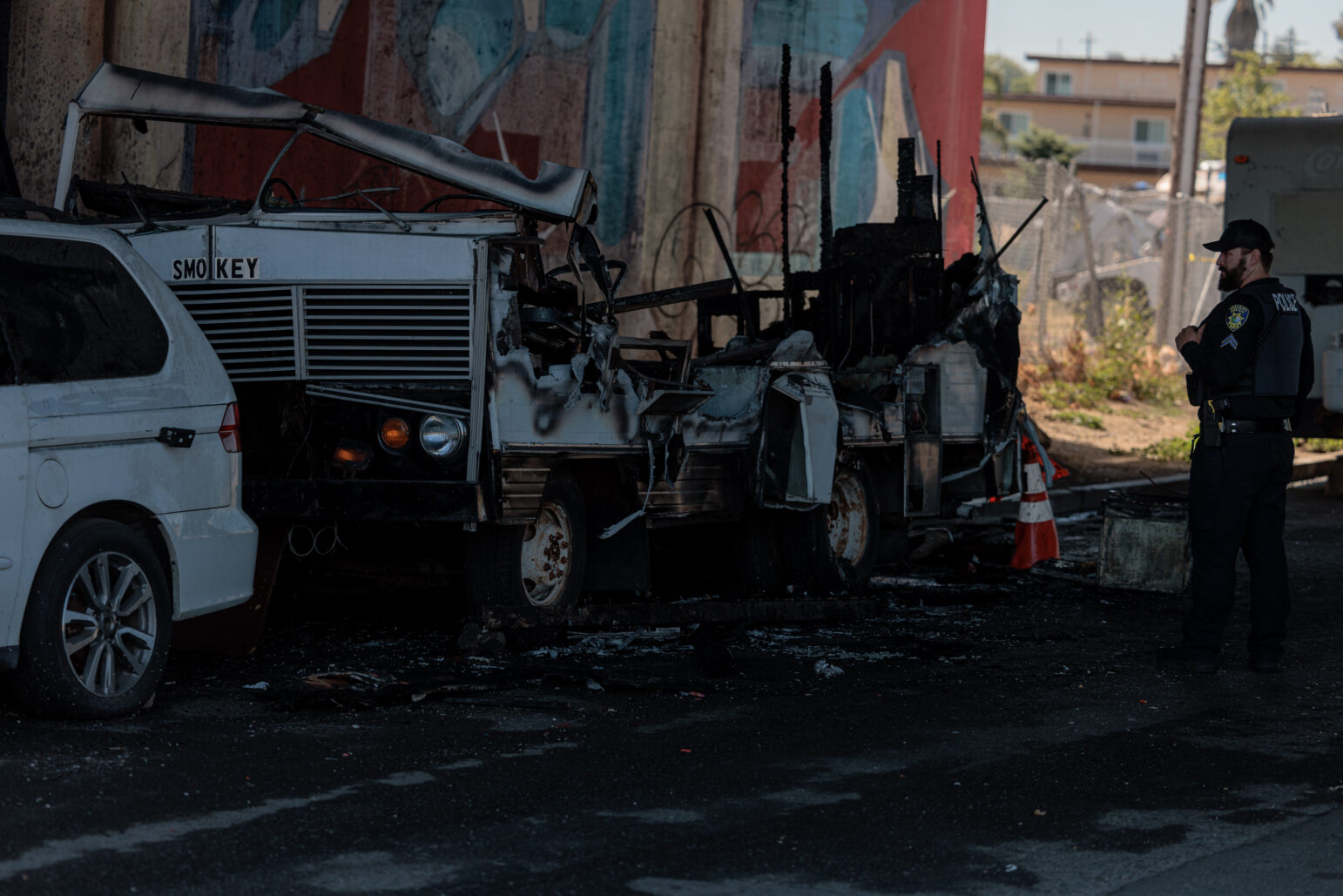 A Vallejo police officer stands near a severely burned RV parked under an overpass. The vehicle, labeled "SMOKEY," is charred and partially collapsed, with debris scattered around it. A white minivan is parked beside the RV, and orange traffic cones are placed near the scene. A colorful mural is partially visible on the concrete pillar in the background.
