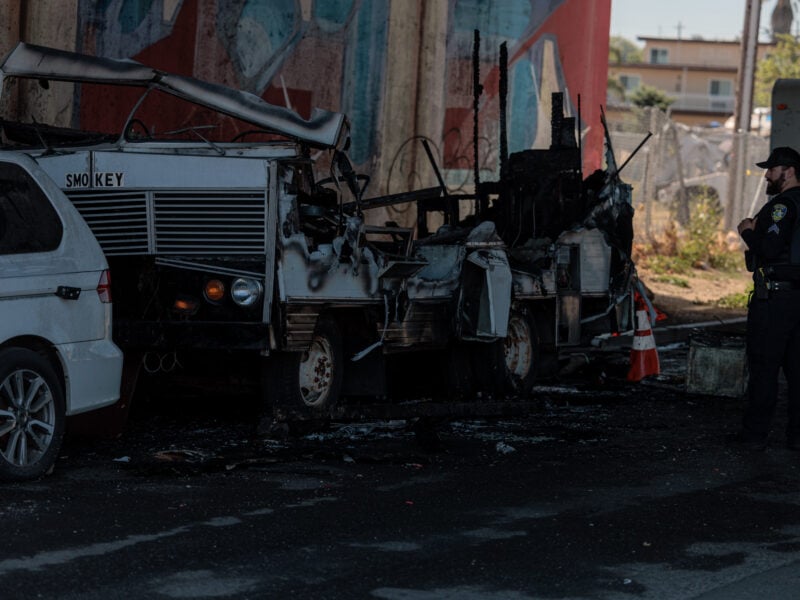 A Vallejo police officer stands near a severely burned RV parked under an overpass. The vehicle, labeled "SMOKEY," is charred and partially collapsed, with debris scattered around it. A white minivan is parked beside the RV, and orange traffic cones are placed near the scene. A colorful mural is partially visible on the concrete pillar in the background.