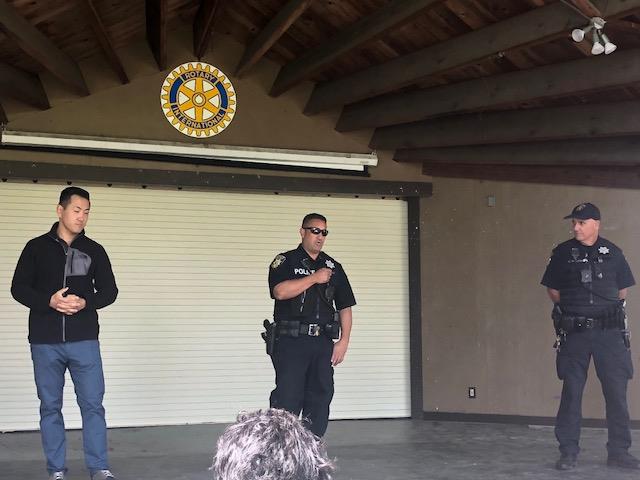 Three police personnel speak to an audience in a small, covered outdoor amphitheater.